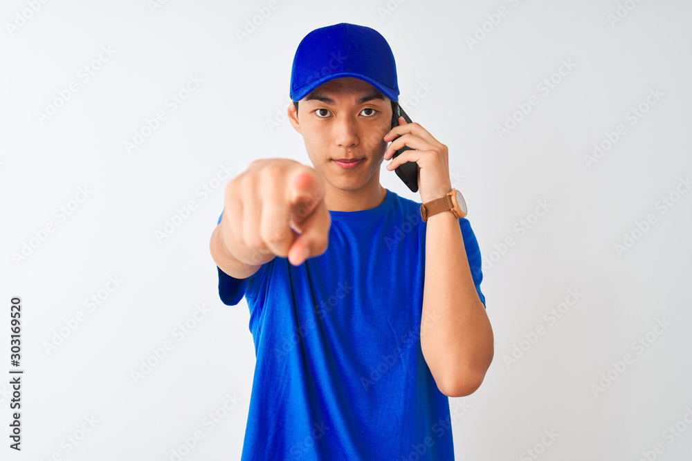 Chinese deliveryman wearing cap talking on the smartphone over isolated white background pointing with finger to the camera and to you, hand sign, positive and confident gesture from the front