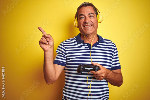 Middle age man playing video game using headphones over isolated yellow background very happy pointing with hand and finger to the side