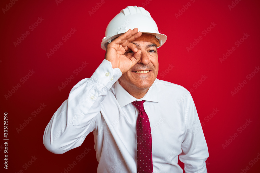 Handsome middle age engineer man wearing security helmet over isolated red background doing ok gesture with hand smiling, eye looking through fingers with happy face.
