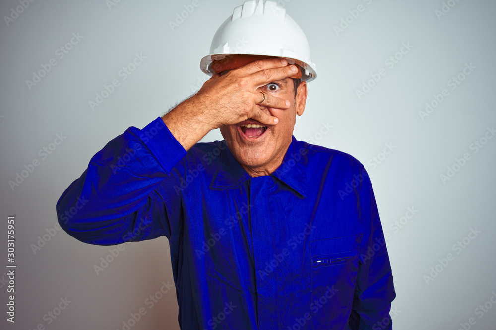 Handsome middle age worker man wearing uniform and helmet over isolated white background peeking in shock covering face and eyes with hand, looking through fingers with embarrassed expression. Handsome middle age worker man wearing uniform and helmet over isolated white background peeking in shock covering face and eyes with hand, looking through fingers with embarrassed expression.