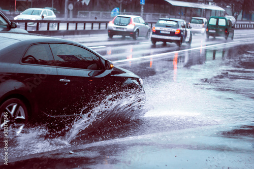Ukraine. Kiev - 05,12,2019 Spraying water from the wheels of a vehicle moving on a wet city asphalt road. The wet wheel of a car moves at a speed along a puddle on a flooded city road during rain. © Yuliia