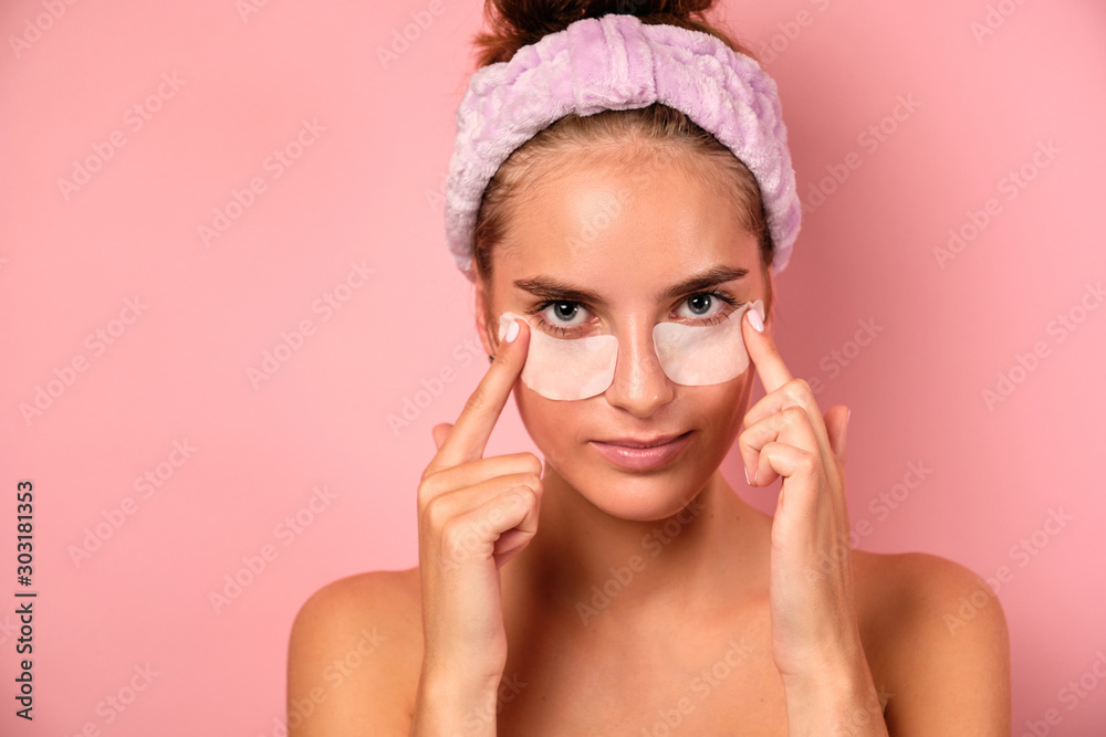A beautiful girl with a bandage on her head holds patches under her eyes while standing on a pink background. A beautiful girl with a bandage on her head holds patches under her eyes while standing on a pink background.