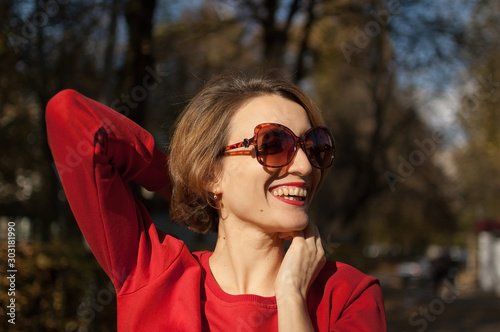 Beautiful smiling woman wearing a sunglasses and red sweater is posing in autumn park during sunny weather outdoors