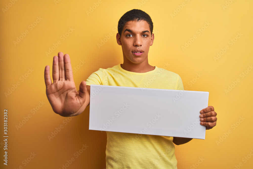 Young handsome arab man holding banner standing over isolated yellow background with open hand doing stop sign with serious and confident expression, defense gesture Young handsome arab man holding banner standing over isolated yellow background with open hand doing stop sign with serious and confident expression, defense gesture