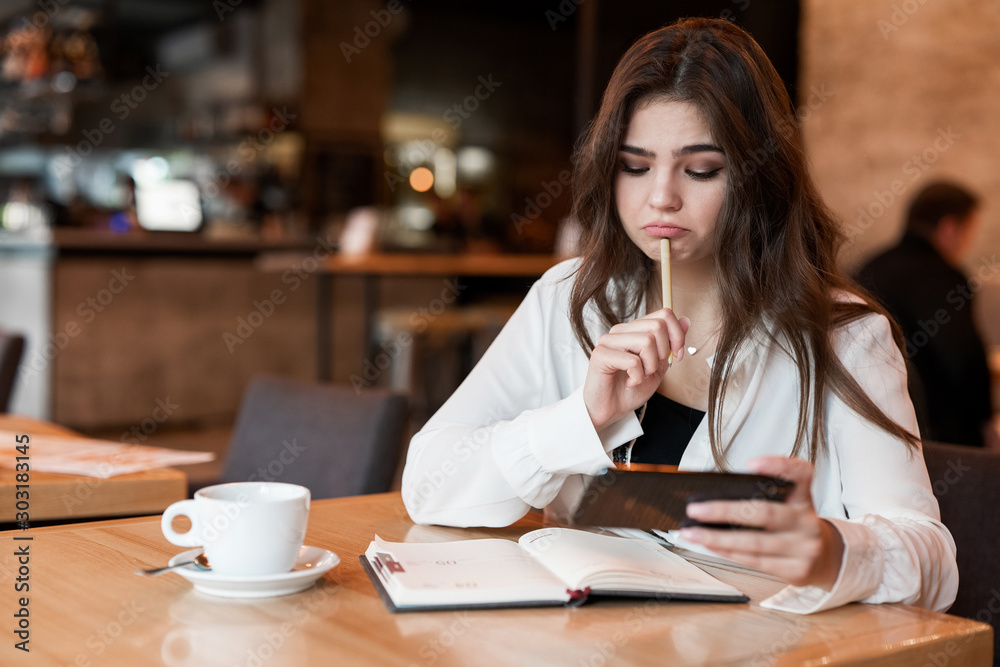 young beautiful woman with pen in her hand taking notes to her planner using her smartphone looks hesitant drinking hot coffee in cafe modern businesswoman young beautiful woman with pen in her hand taking notes to her planner using her smartphone looks hesitant drinking hot coffee in cafe modern businesswoman