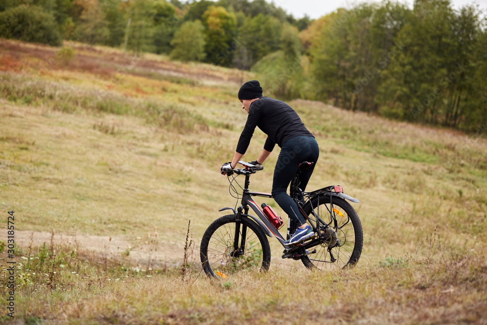 Image of mauntain bike cyclist in meadow near forest, side view of sporty male riding bike during his vacation, enjoying his hobby, rides downhill, covering assigned distance. Lifestyle concept.