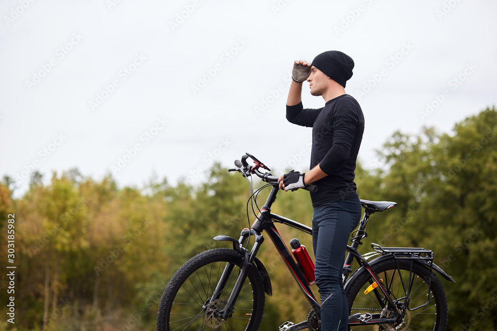 Image of road biker cycling and training on road in forest. Attractive young sportsman stops to have rest after long hours riding, keeps hand near forehead and looks faraway. Healthy activity concept.