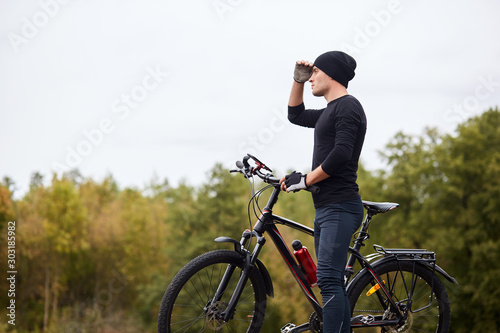 Fototapeta Naklejka Na Ścianę i Meble -  Image of road biker cycling and training on road in forest. Attractive young sportsman stops to have rest after long hours riding, keeps hand near forehead and looks faraway. Healthy activity concept.