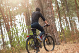 Back view of biker on trail in woods, young man cycling MTB in forest, handsome man wearing black track suit spending time in open air, male riding uphill. Sport, fitness, motivation concept.
