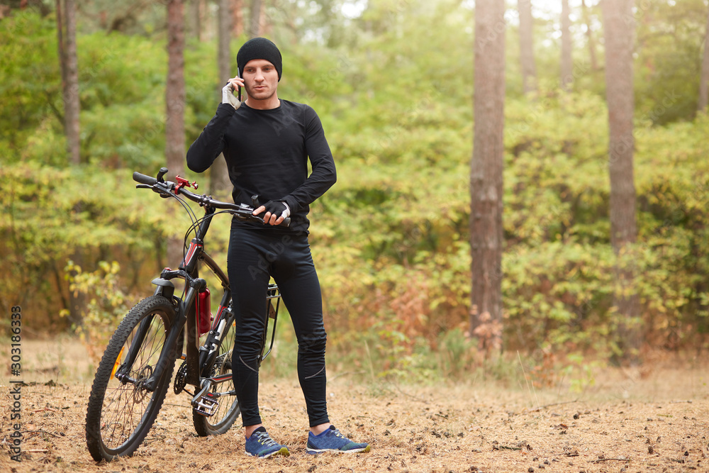 Obraz premium Outdoor shot of man wearing black track suit and sneakers, standing near his mountain bike and speaks phone with beautiful forest on backgraund, spending time in active way. Healthy lifestyle concept.