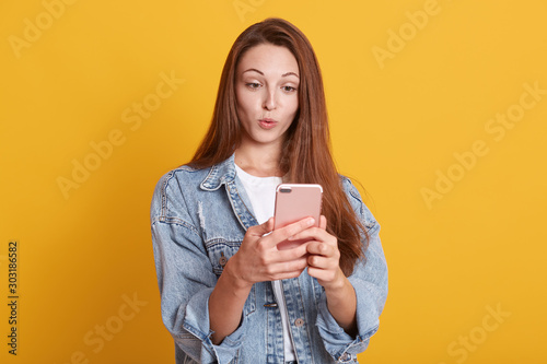 Portrait of attractive Cauacasian woman gets surprising news via telephone call, keeps lips rounded, wears stylish denim jacket, has eyes popped out, hears astonishing relevation, standing indoor.