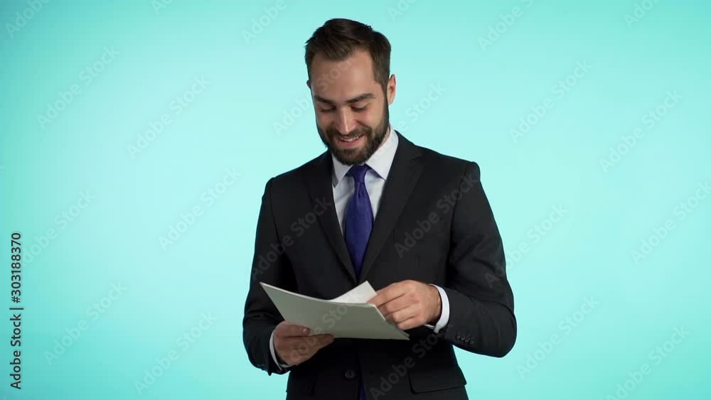 Young handsome man in suit jacket checks documents, utility bills, report. Businessman isolated on blue background. He is satisfied with work of corporate staff. Young handsome man in suit jacket checks documents, utility bills, report. Businessman isolated on blue background. He is satisfied with work of corporate staff.
