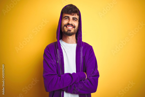 Young man with tattoo wearing purple sweatshirt with hood over isolated yellow background happy face smiling with crossed arms looking at the camera. Positive person.