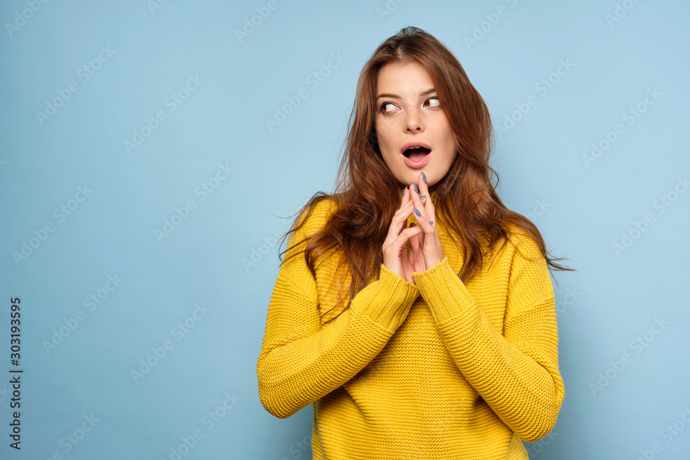 The brunette stands on a blue background in a yellow sweater and looks away, opening her eyes and mouth wide The brunette stands on a blue background in a yellow sweater and looks away, opening her eyes and mouth wide