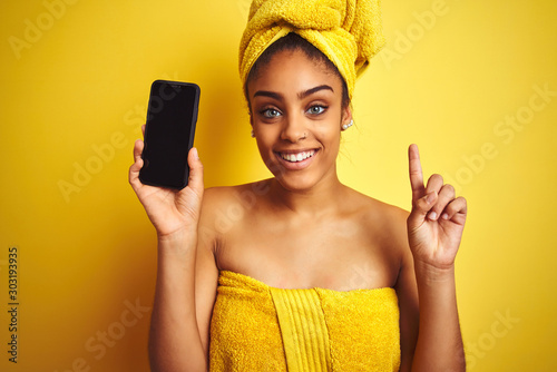Afro woman wearing towel after shower showing smatrphone over isolated yellow background surprised with an idea or question pointing finger with happy face, number one