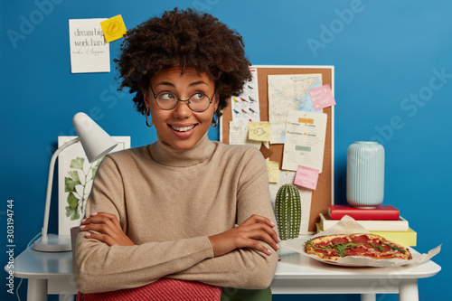 Horizontal shot of cheerful curly haired young woman keeps hands crossed, looks away, smiles pleasantly, dressed casually, wears transparent glasses, poses against blue background with desktop