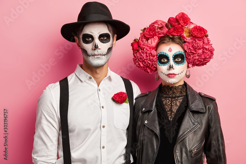 Horizontal shot of serious woman and man dressed in Halloween costumes, wear skeleton makeup, wreath made of peonies, enjoy photoshoot, wait for party or carnival. Dead couple during All Souls Day