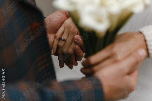 Groom holding hands of the bride with the ring on her finger and wedding bouquet
