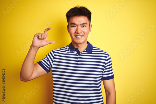 Young asian chinese man wearing striped polo standing over isolated yellow background smiling and confident gesturing with hand doing small size sign with fingers looking and the camera