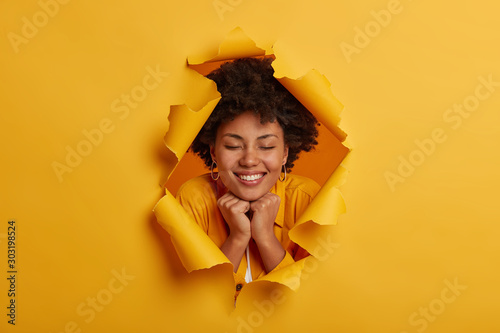 Pleased curly haired woman keeps hands under chin, smiles broadly, has eyes closed, looks through paper hole, makes creative shot, expresses happy emotions, imagines pleasant thing. Ripped background © wayhome.studio 