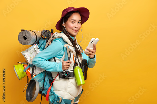 Pretty satisfied traveler uses free internet connection on smartphone for blogging during wanderlust trip, carries big heavy rucksack, has binoculars and retro camera to explore surroundings