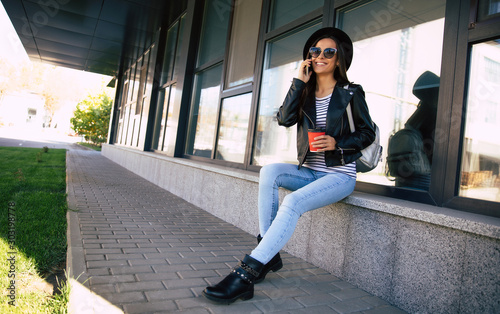 Coffee break. Gorgeous woman in casual outfit is sitting on a bench and talking on the phone while holding a red coffee cup, laughing and enjoying herself.