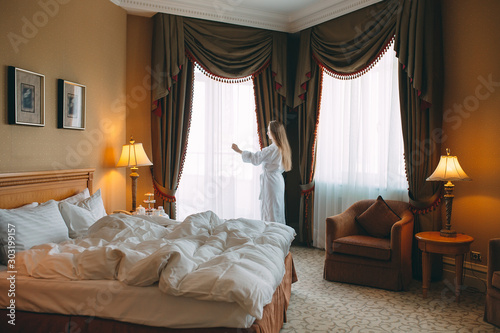 Woman in bathrobe stay near the window in hotel room.