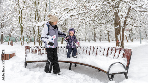 Family walks during snowfall in Moscow, Russia. Baby girl with mother are in winter park. Little child walks on snowbound bench by the mom's hand. Young woman and her kid play with snow outdoor. photo