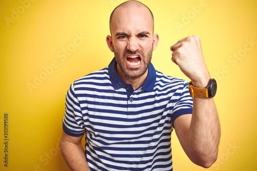 Young bald man with beard wearing casual striped blue t-shirt over yellow isolated background angry and mad raising fist frustrated and furious while shouting with anger. Rage and aggressive concept.