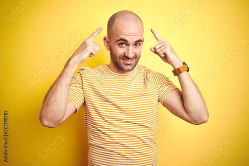 Young bald man with beard wearing casual striped t-shirt over yellow isolated background smiling pointing to head with both hands finger, great idea or thought, good memory