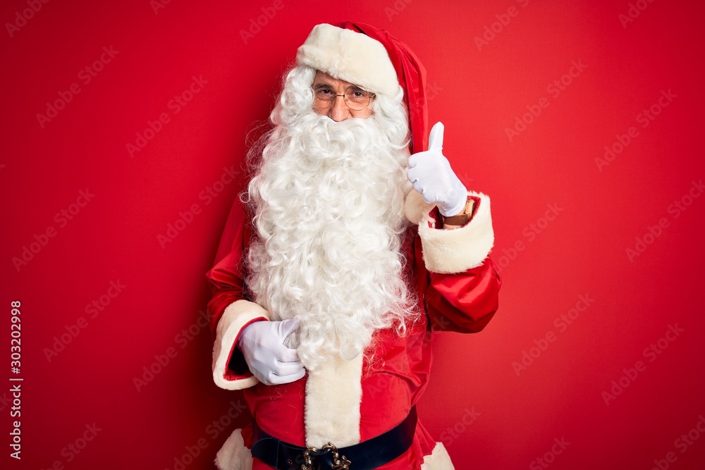 Middle age handsome man wearing Santa costume standing over isolated red background doing happy thumbs up gesture with hand. Approving expression looking at the camera showing success.