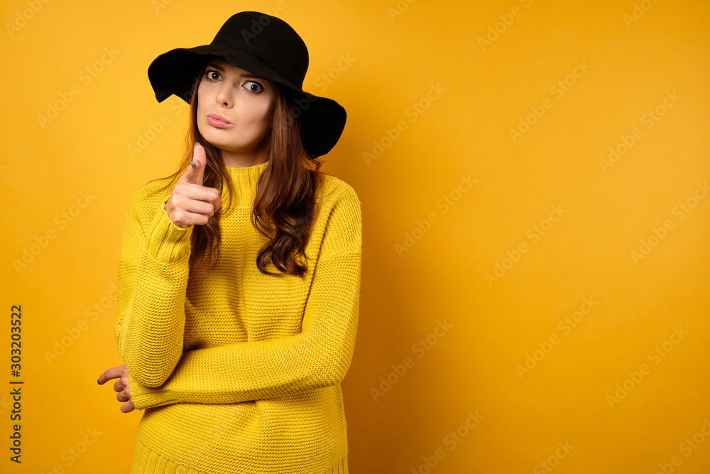 A beautiful brunette in a yellow sweater and black hat stands on a yellow background frowning and pointing her finger at the frame A beautiful brunette in a yellow sweater and black hat stands on a yellow background frowning and pointing her finger at the frame