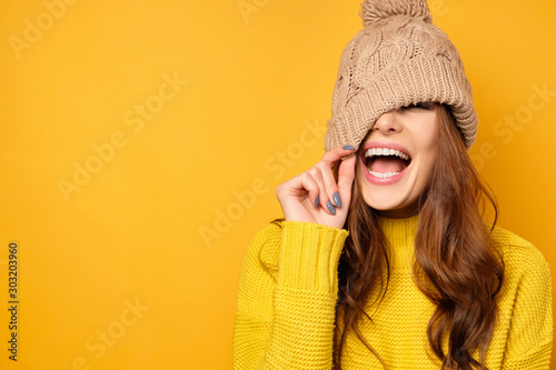 A brunette stands on a yellow background in a yellow sweater, pulls the hat down and laughs joyfully