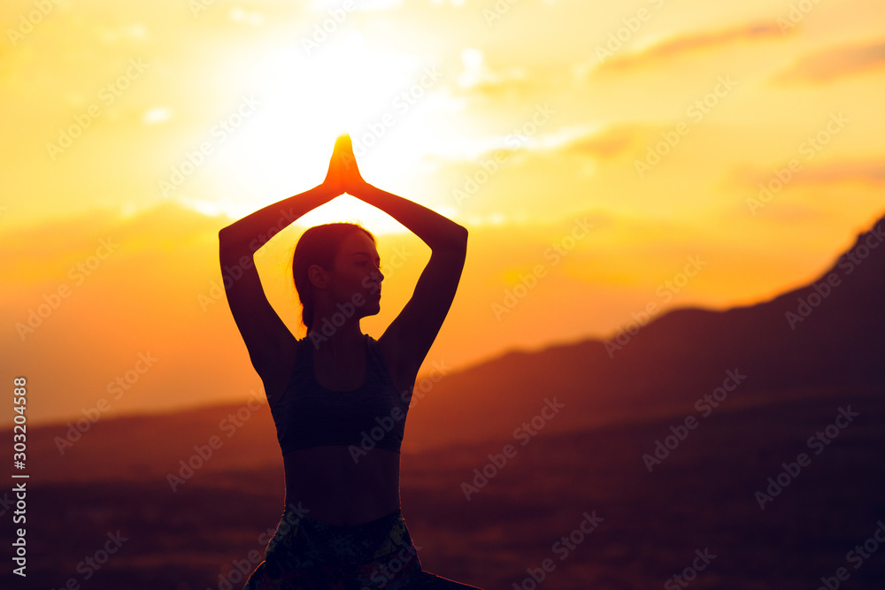 Silhouette of young woman practicing yoga or pilates at sunset or sunrise in beautiful mountain location.