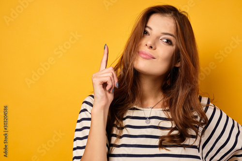 A beautiful brunette in a striped T-shirt stands on a yellow background holding her index finger up.