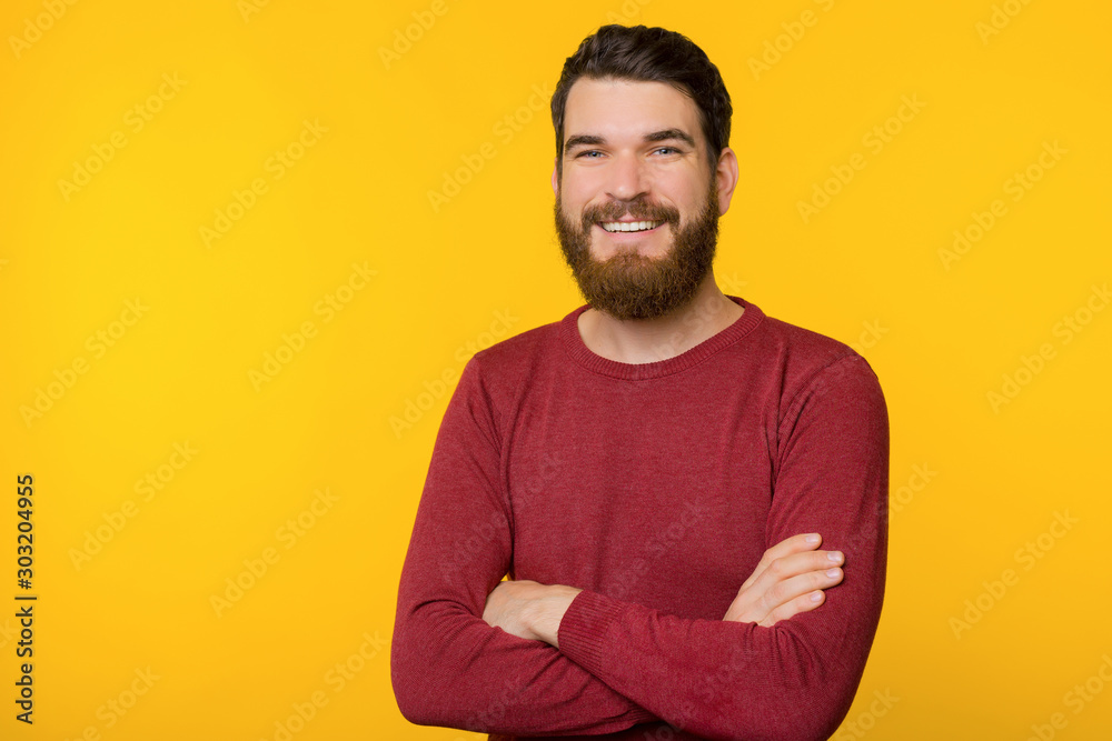 Photo of bearded guy, standing with crossed arms, looking and smiling at camera  over yellow isolated background - obrazy, fototapety, plakaty 