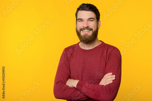 Photo of bearded guy, standing with crossed arms, looking and smiling at camera  over yellow isolated background