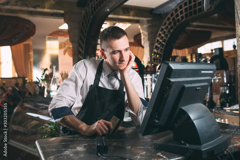 small business, people and service concept - happy man or waiter in apron at counter with cashbox working at bar or coffee shop small business, people and service concept - happy man or waiter in apron at counter with cashbox working at bar or coffee shop