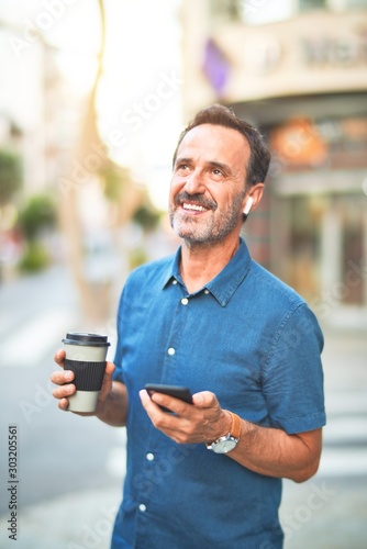 Middle age handsome businessman standing on the street using smartphone and earphones drinking coffee