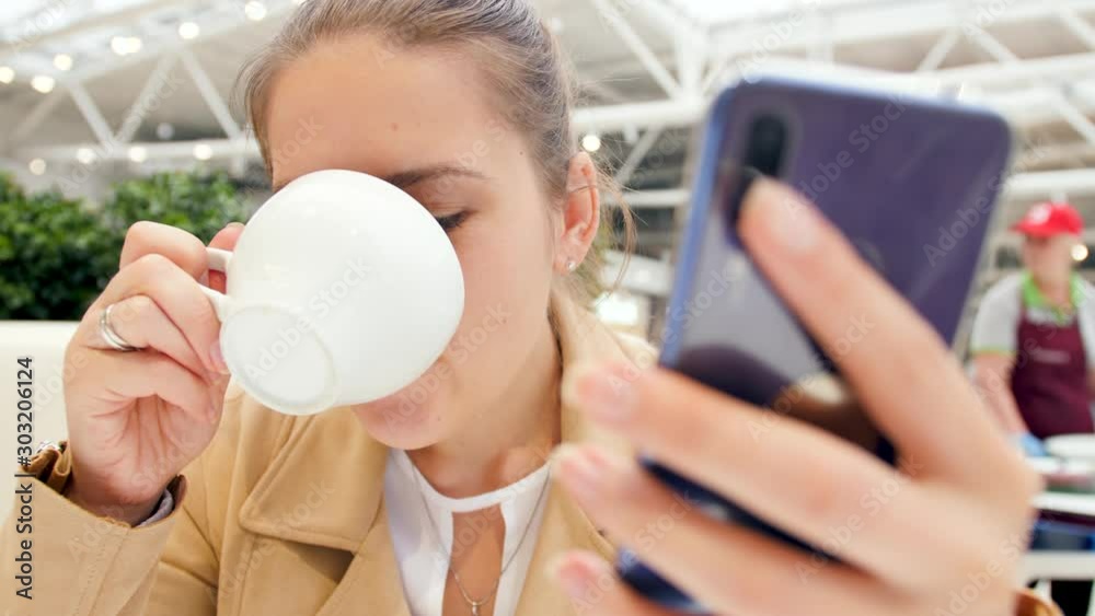 Closeup 4k video of young woman holding smartphone and typing message while drinking cappuccino coffee in cafeteria