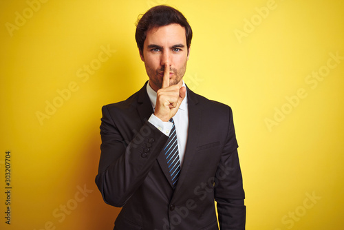 Young handsome businessman wearing suit and tie standing over isolated yellow background asking to be quiet with finger on lips. Silence and secret concept.