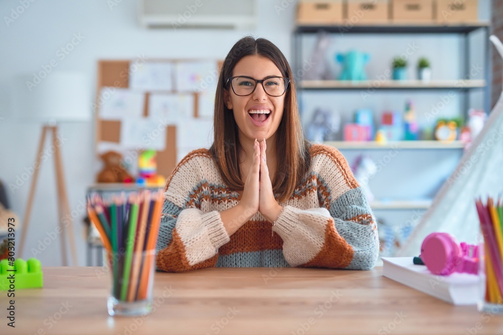 Young beautiful teacher woman wearing sweater and glasses sitting on desk at kindergarten praying with hands together asking for forgiveness smiling confident. Young beautiful teacher woman wearing sweater and glasses sitting on desk at kindergarten praying with hands together asking for forgiveness smiling confident.