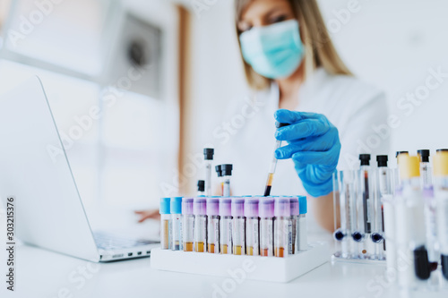 Close up of lab assistant in uniform, with mask and rubber gloves holding test tube with blood sample while sitting on chair and typing on laptop. Selective focus on test tubes. photo