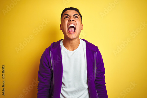 Young brazilian man wearing purple sweatshirt standing over isolated yellow background angry and mad screaming frustrated and furious, shouting with anger. Rage and aggressive concept.