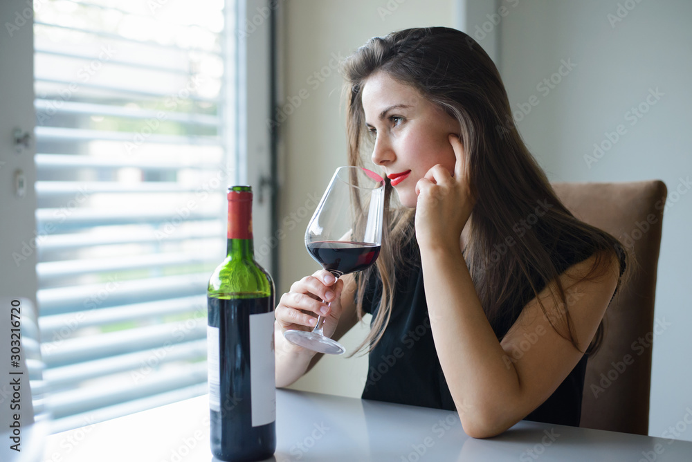 Lonely girl sitting in kitchen drinking red sweet wine. Beautiful caucasian female looking through the window drinking alcohol dreaming of someone Lonely girl sitting in kitchen drinking red sweet wine. Beautiful caucasian female looking through the window drinking alcohol dreaming of someone