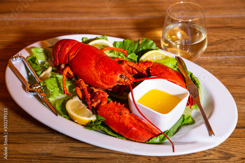 Boiled lobster served on a white ceramic platter with lemon slices and romaine lettuce, melted butter, and white wine in a stemless wine glass, against a wood background.