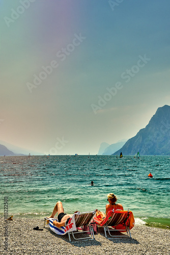 Riva del Garda,Lago di Garda ,Italy - 21 June 2018: People who bathe and sit on the beach to tan at Lake Garda, beautiful Lake Garda surrounded by mountains in the summer time
