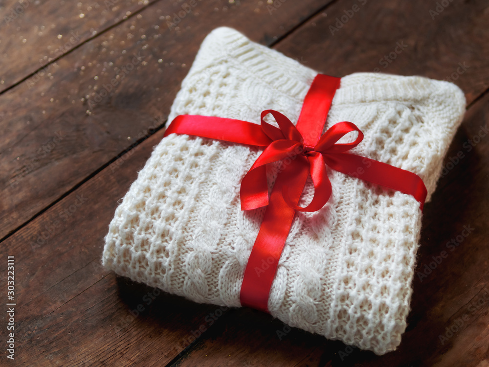 White  knitted sweater with red ribbon bowknot on shabby wooden background. Cozy present for holiday.