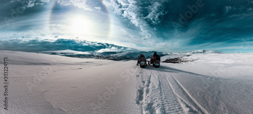 Two snowmobiles with one driver stand in front of dramatic scenic view at cold snowy Norwegian Mountains, Sun halo shines on cold skies, dark blue clouds flying from Atlantic Ocean. Lappland, Sweden