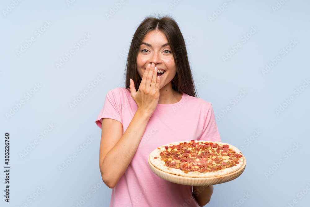 Pretty young girl holding a pizza over isolated blue wall with surprise facial expression